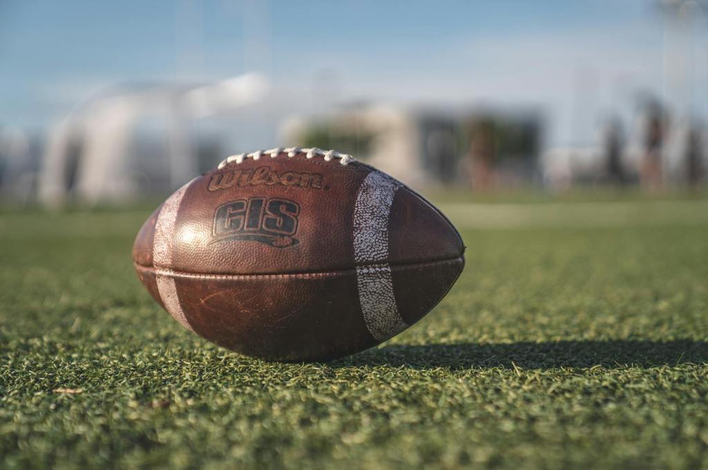A worn football sits on a turf field in an outdoor setting.