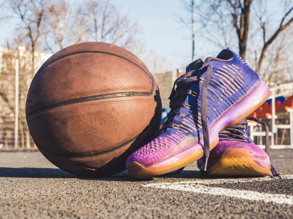 A well-worn basketball sits next to a pair of purple and magenta sneakers on an outdoor court.