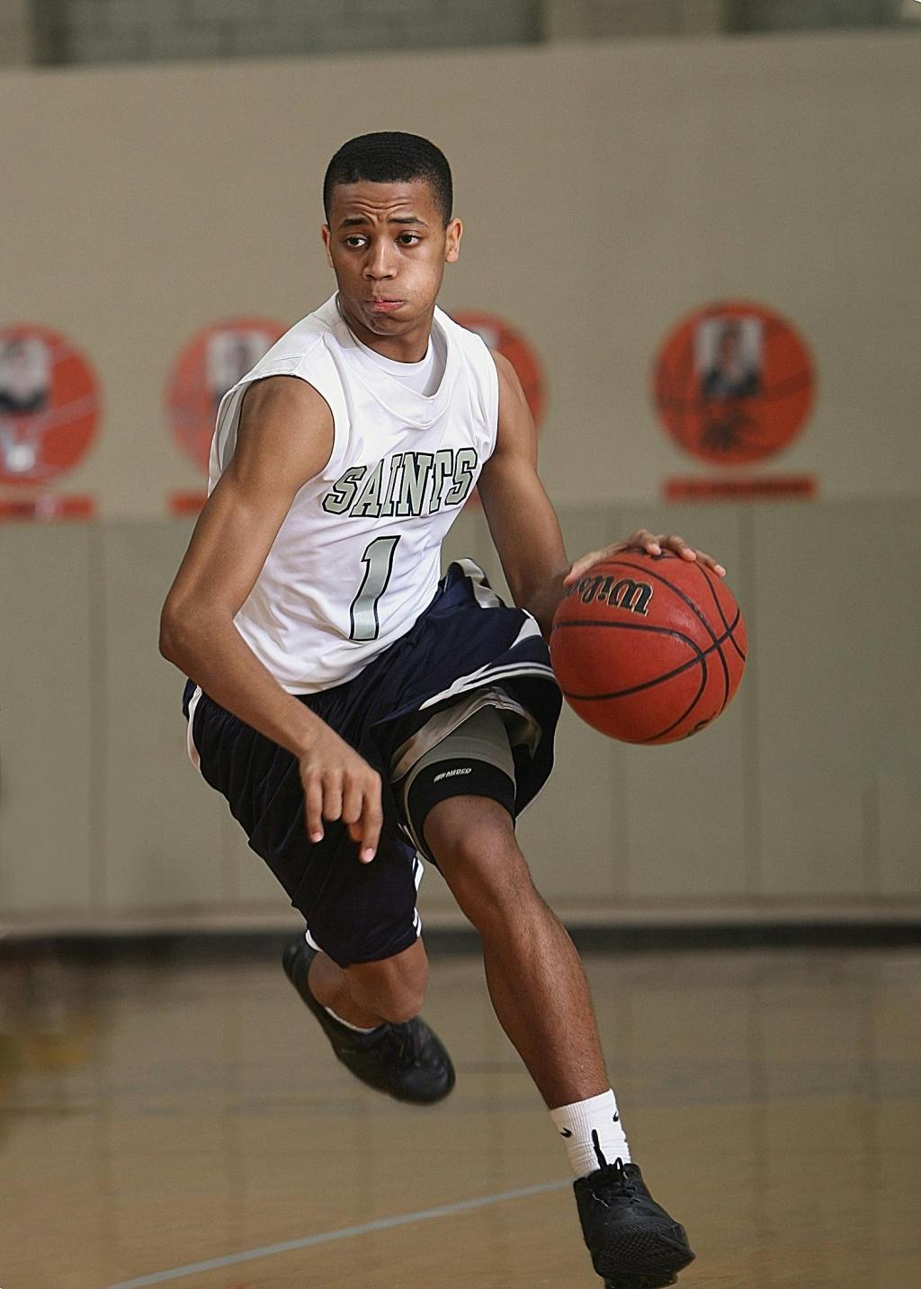 A male, African American basketball player dribbles a ball while running on an indoor basketball court. His face is focused with concentration.