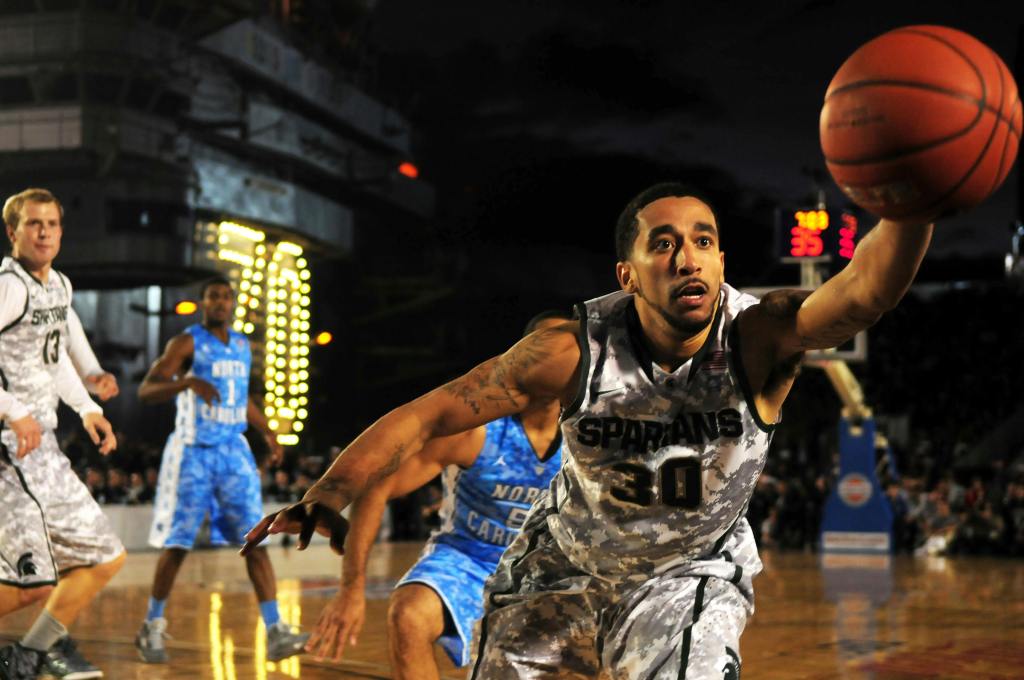 An African American basketball player dives for a basketball in an indoor arena while other players approach from behind.